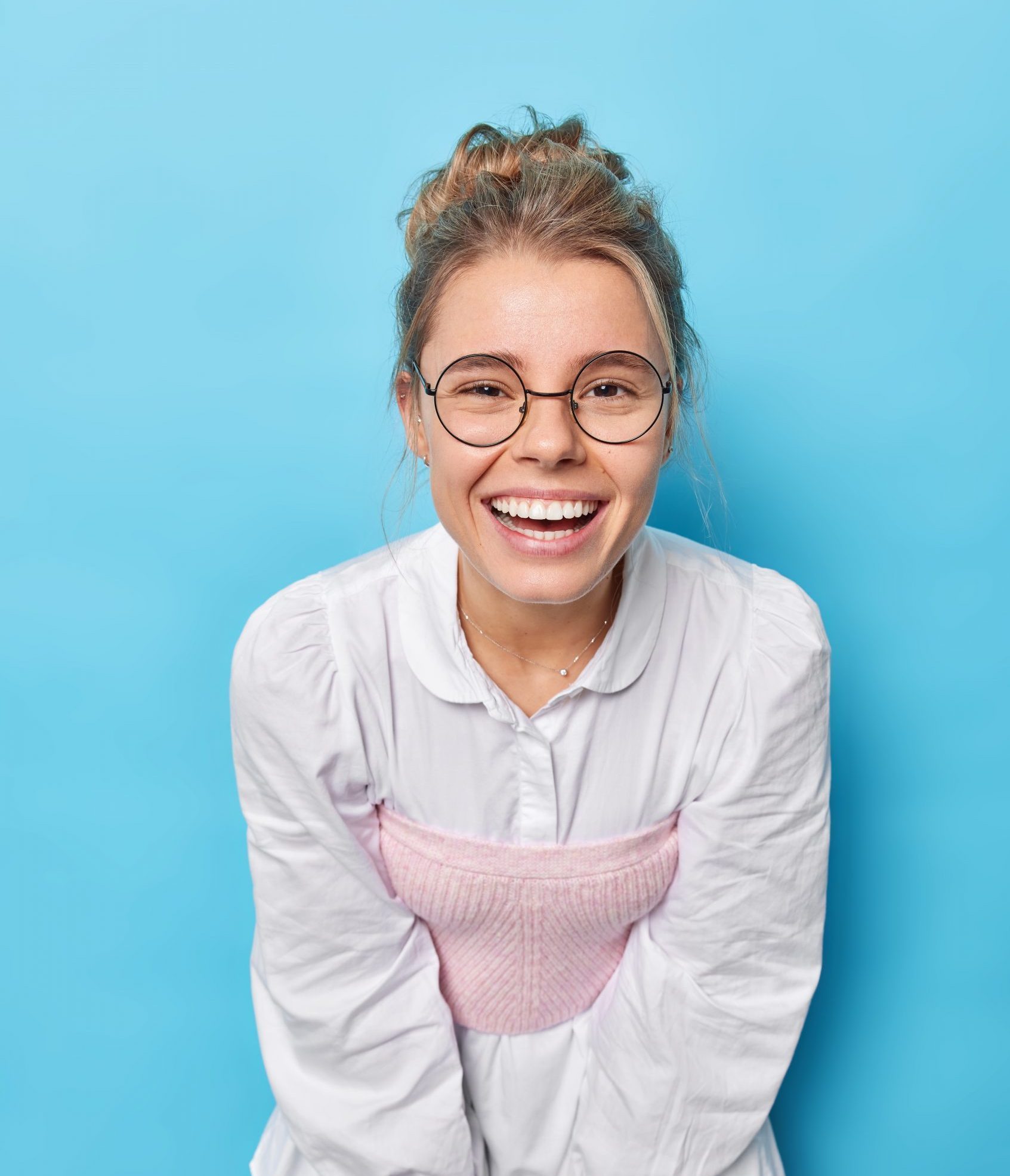Vertical shot of cheerful young European woman dressed in formal white blouse wears round spectacles leans forward has upbeat mood isolated over blue studio background. Happy emotions concept